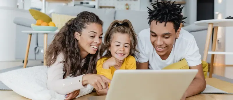 A smiling family of three, including a mother, father, and young daughter, is lying on the floor looking at a laptop together.
